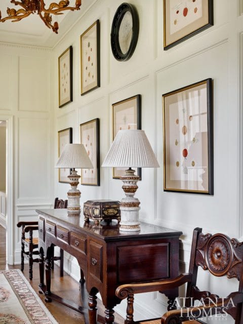 Hallway with paneled walls, a dark wood console table with two lamps and a decorative box, framed botanical prints, and ornate wooden chairs.
