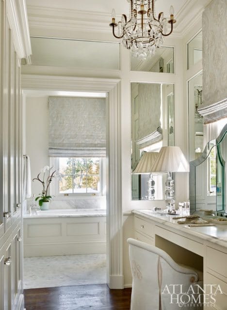 White dressing room with a vanity, upholstered chair, mirrored walls, and a crystal chandelier. A window with a roman shade looks into a marble-clad bathroom.