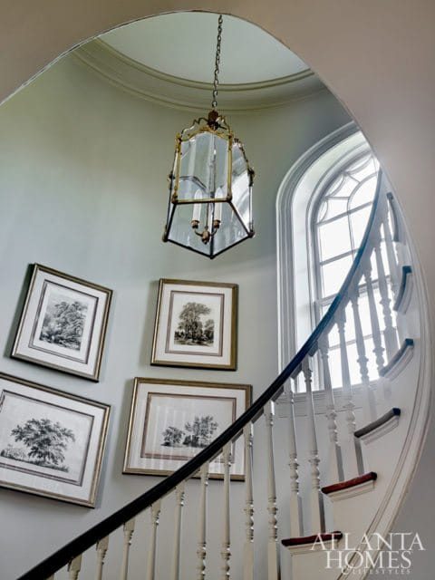 Curved staircase with white balusters and dark handrail, adorned with four framed botanical prints and a brass and glass lantern chandelier.