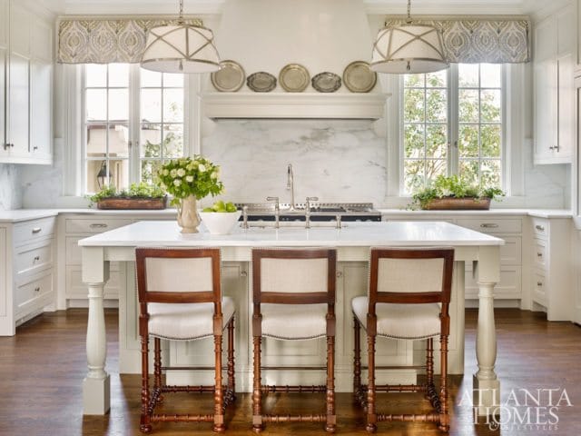 White kitchen with marble backsplash, large island with three upholstered stools, and two pendant lights.