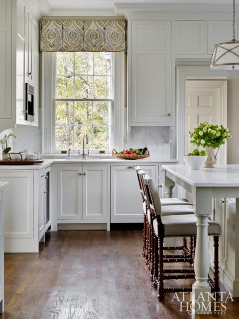White kitchen with marble countertops, paneled cabinets, and a large window overlooking trees. Bar stools with dark wood frames and upholstered seats are at a marble-topped island.