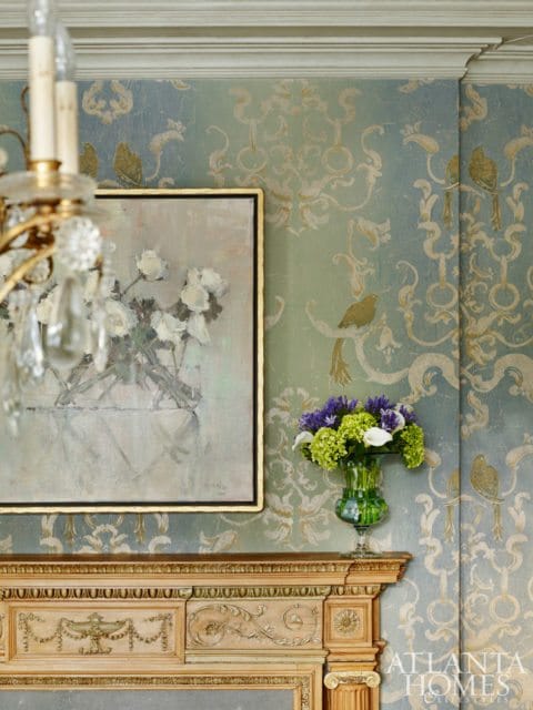 Ornate wooden mantelpiece below a painting of white roses and a vase of purple and green flowers, set against blue and green damask wallpaper.