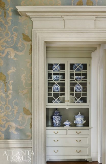 Built-in light gray cabinet with glass doors displaying blue and white porcelain dishes, and drawers with brass hardware. Blue and white ginger jars and a teapot sit on top of the drawers.