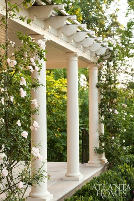 White pergola with decorative trim supported by fluted columns, adorned with climbing pink roses. Lush greenery and trees in the background.