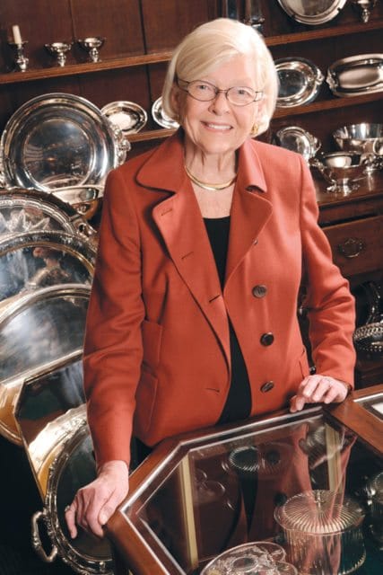 Elderly woman with blonde hair and glasses wearing a rust-colored blazer and black top, standing in front of shelves filled with silver serving dishes and bowls.