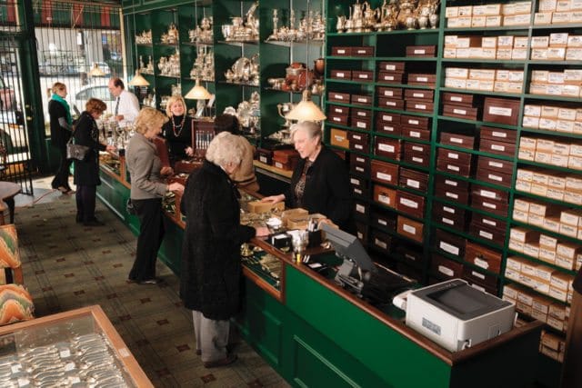 Interior of a shop with green shelving displaying silver items and numerous wooden boxes. Customers interact with staff at a green counter.