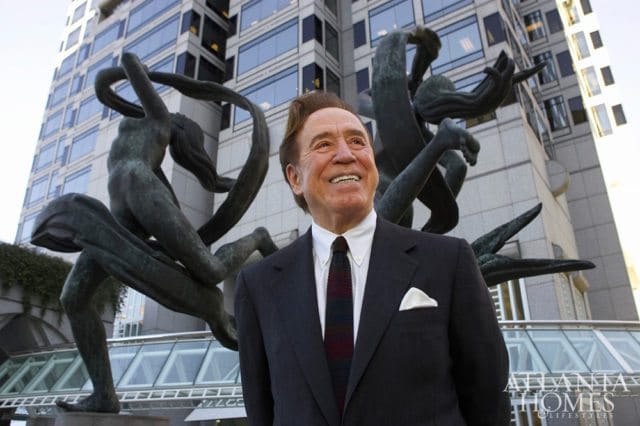 Man in a dark suit and tie smiles while standing in front of a building with abstract sculptures.