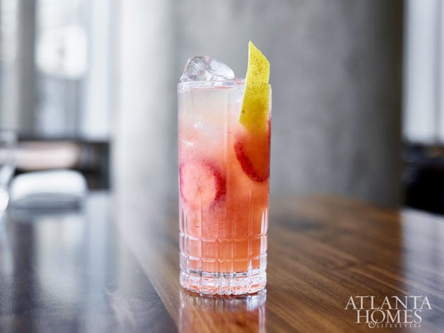 Tall glass of pink cocktail with ice, strawberry slices, and a pear slice garnish, on a wooden table.