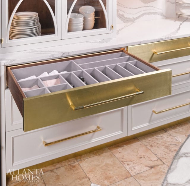 White kitchen cabinetry with brass hardware and a marble countertop. An open drawer reveals a gray-lined organizer with compartments.