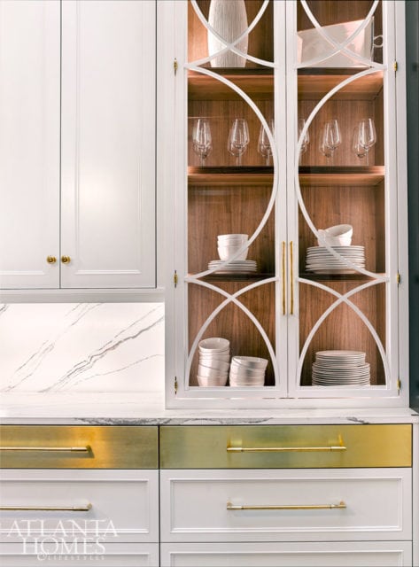 White kitchen cabinetry with glass-fronted display cabinets featuring stacked white bowls and plates, and brass hardware on gold and white drawers.