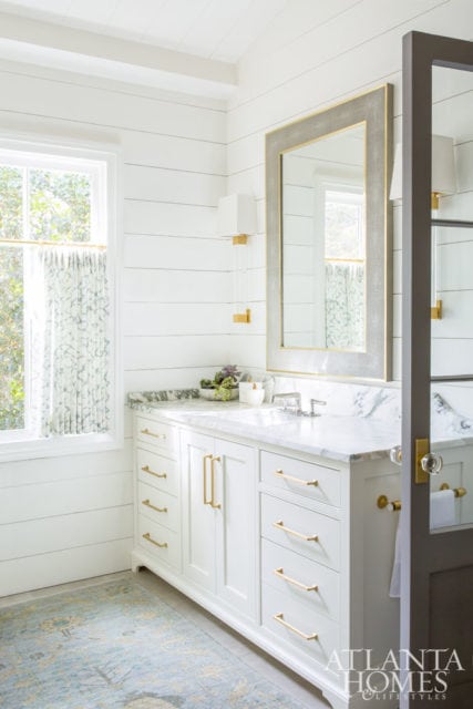 White bathroom vanity with marble countertop, brass hardware, and shiplap walls. A large framed mirror hangs above the vanity, and a patterned rug lies on the floor.