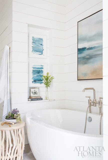 White bathroom with shiplap walls, freestanding soaking tub, abstract blue artwork, and nickel fixtures.