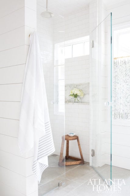 White subway tile shower with glass enclosure, shiplap accent wall, wooden stool, and marble niche with hydrangeas.