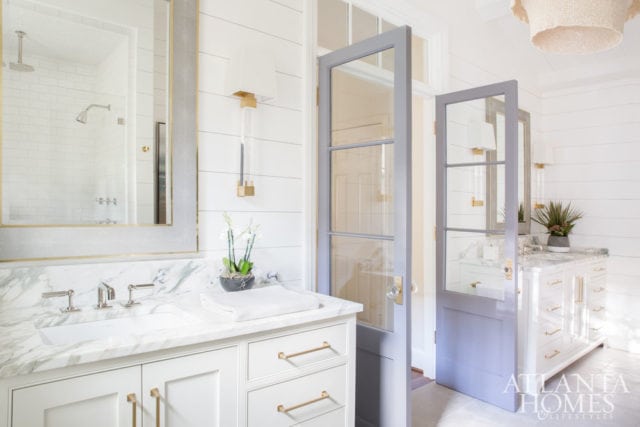 White bathroom with marble countertops, double vanity, brass hardware, and shiplap walls. A gray door opens to another vanity area.