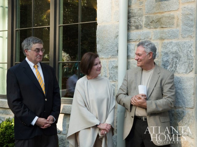 Three people stand outside a stone building. A man in a dark suit and yellow tie stands on the left. A woman in a cream-colored cape stands in the center. A man in a tan jacket stands on the right, holding a white paper. The Atlanta Homes & Lifestyles logo is visible on the right side of the image.