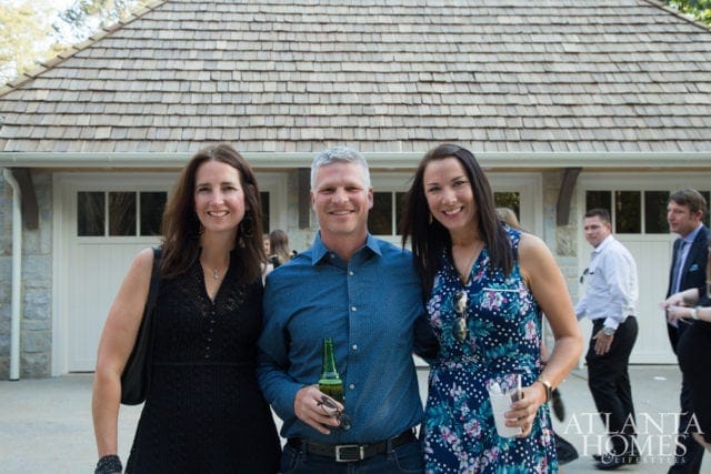 Three people smiling in front of a building with a shingled roof and stone accents. The person on the left wears a black dress, the person in the middle wears a blue patterned shirt and holds a beer, and the person on the right wears a floral dress.