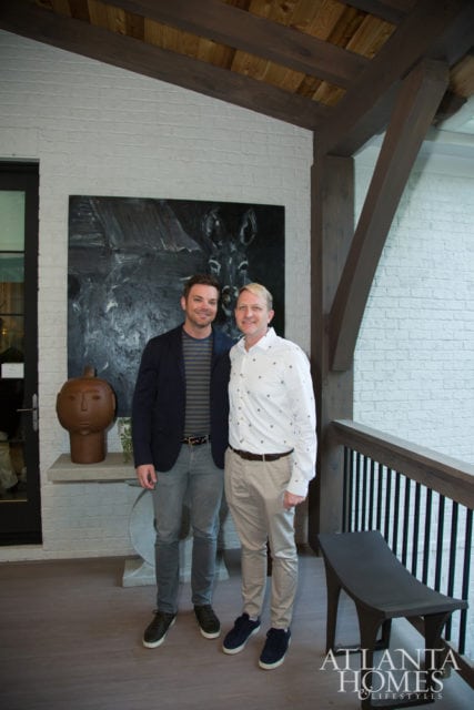 Two men stand on a covered porch with white brick walls and exposed wooden beams. A large abstract painting hangs behind them.