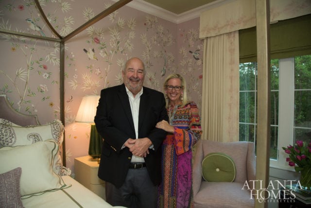 A man in a black blazer and a woman in a colorful dress stand in a bedroom with floral wallpaper and a four-poster bed.