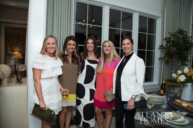 Five women in dresses and a white blazer pose for a photo in a well-decorated room with large windows and a table set for dining.