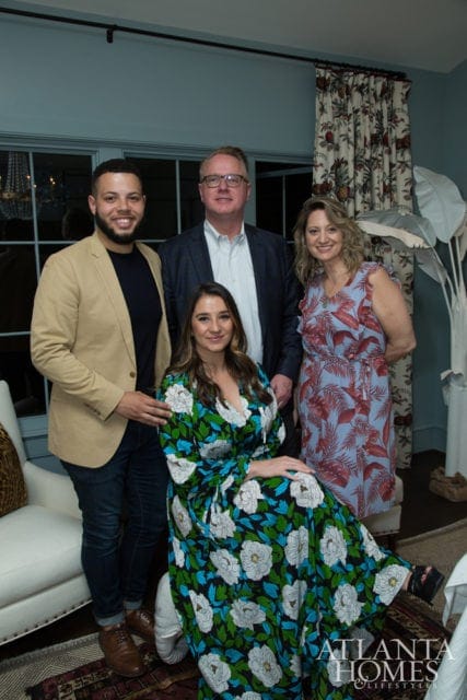 Four people pose for a photo in a room with blue walls and patterned curtains. One woman in a floral dress sits in the foreground.