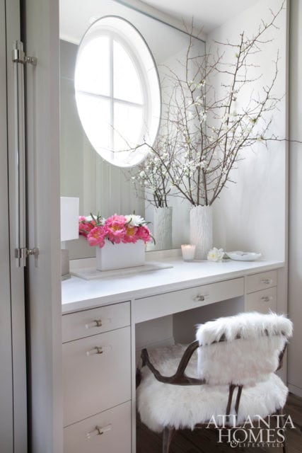 White vanity with crystal knobs, a round window above, and a furry white chair. A vase of pink peonies sits on the vanity.