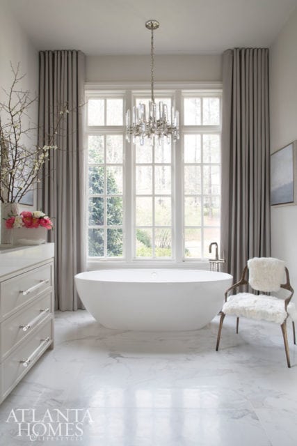 Modern white bathroom with freestanding tub, crystal chandelier, marble floors, and a chair with white fur cushion.