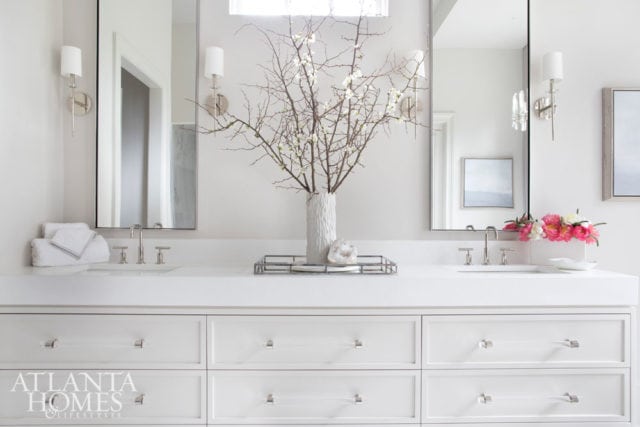 White bathroom vanity with double sinks, chrome faucets, and clear acrylic drawer pulls. A white vase with branches and a geode sit on the counter, flanked by sconces and mirrors.