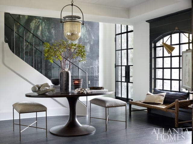 Foyer with a dark wood tulip table, two upholstered stools, and a large abstract mural behind a staircase. A large glass pendant light hangs above the table.