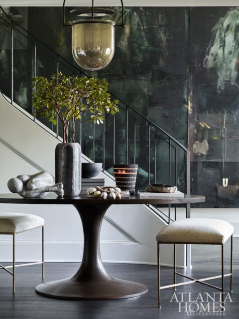 Round dark wood table with sculptural base, topped with a vase of greenery, a sculpture, and decorative objects. Two upholstered stools with thin metal legs flank the table. A large pendant light hangs above.