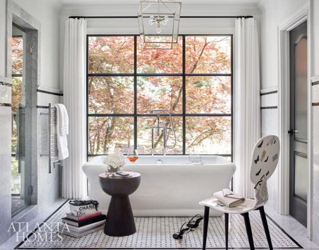 White freestanding bathtub in a bathroom with a large window overlooking trees with fall foliage. Black grid window frame, white tile floor, marble accents, and a modern lantern light fixture.