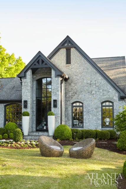 Stone-clad house with dark trim, tall arched windows, and a prominent gable. Two woven chairs sit on the lawn in front of manicured hedges and white planters.