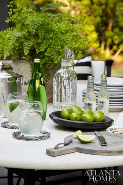 Outdoor table setting with drinks, limes, and a fern, featuring a Perrier bottle and two glasses with ice and lime wedges.
