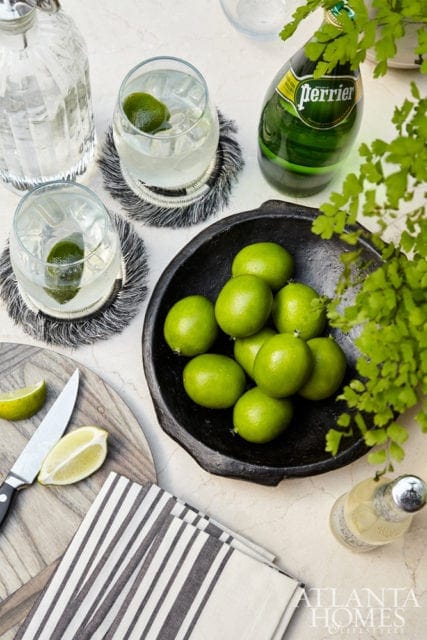 A black bowl filled with fresh limes sits next to two glasses of ice water with lime slices, a bottle of Perrier, and a knife with lime wedges on a cutting board.