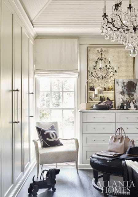 Creamy white dressing room with floor-to-ceiling cabinetry, a tufted armchair with a Chanel pillow, a crystal chandelier, and a dresser topped with accessories.