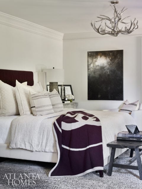 Bedroom with a white bed, dark upholstered headboard, and a plum throw blanket with white stripes. A silver antler chandelier hangs above.