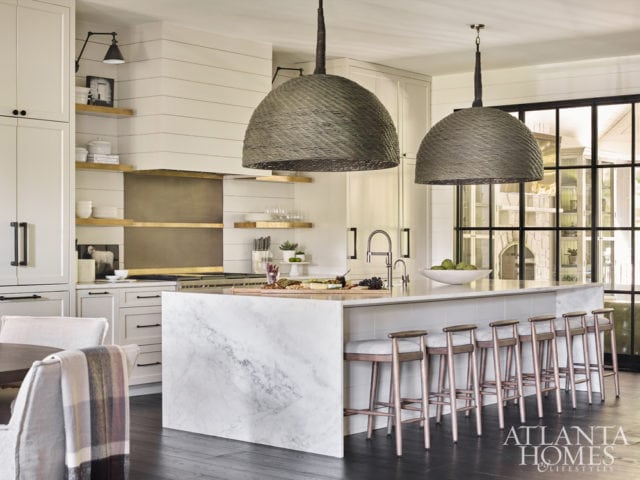 Modern kitchen with white shiplap range hood, marble island with bar stools, and large woven pendant lights. A large black grid window is visible in the background.