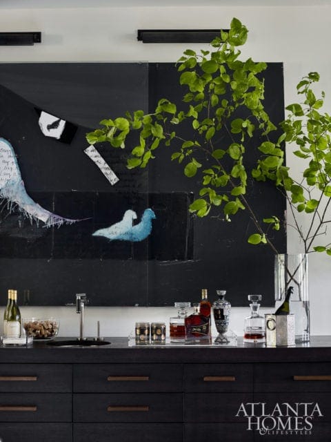 Dark wood bar with bottles of liquor and wine, a sink, and a large abstract black and white painting with green branches in a vase.