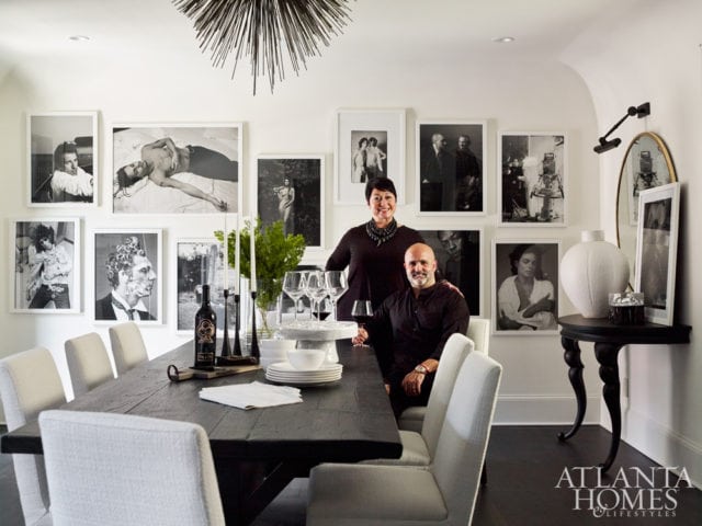 Dining room with dark wood table, white upholstered chairs, and a gallery wall of black and white photographs. A couple stands behind the table.