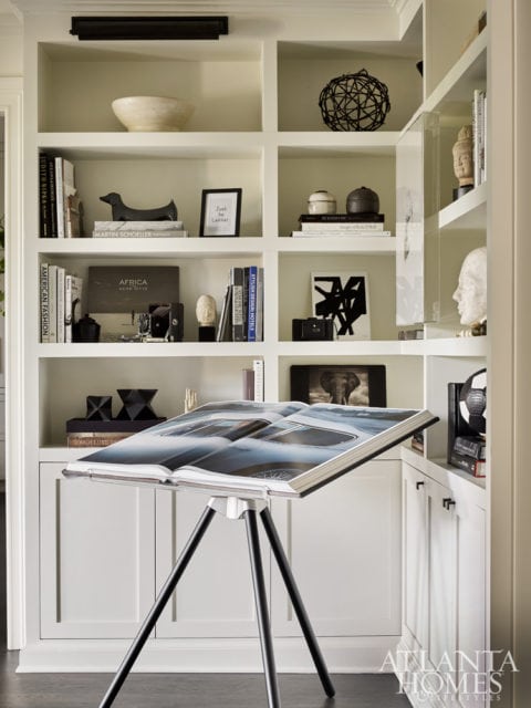 Built-in white shelves display books, decorative objects, and artwork, with an open book on a tripod stand in the foreground.