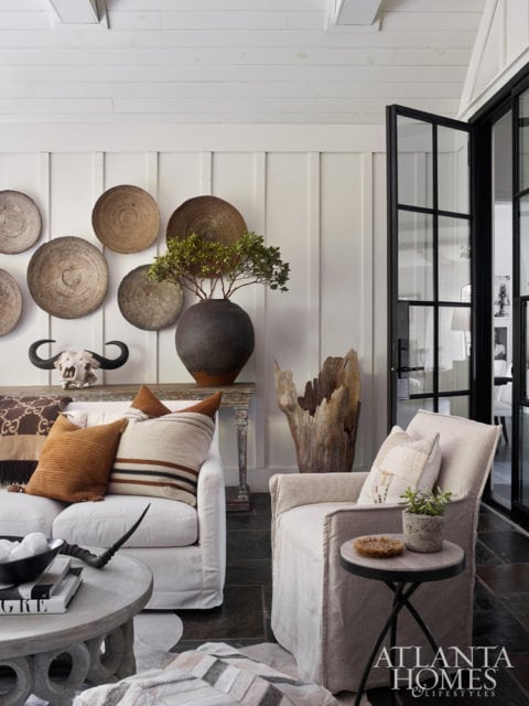 Living room with white sofa and armchair, textured pillows, round coffee table, wall decor of woven baskets and a bull skull, and a large wooden sculpture.