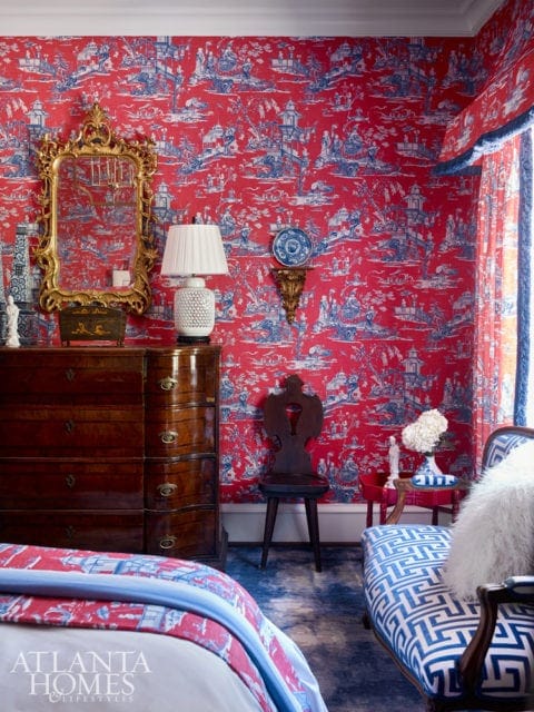 Bedroom with red and blue toile wallpaper, a dark wood dresser, a gold ornate mirror, and a blue and white patterned chaise lounge.