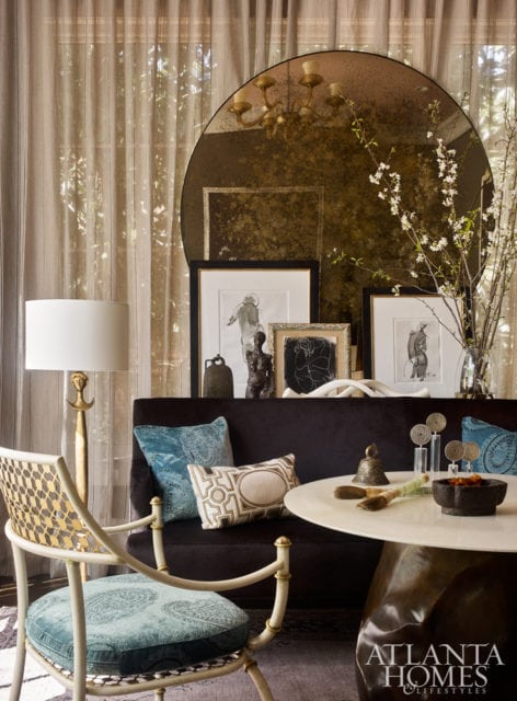 Living room with a dark velvet sofa, a round marble-top table, and a decorative chair with blue paisley cushions. A large, distressed mirror hangs above the sofa.