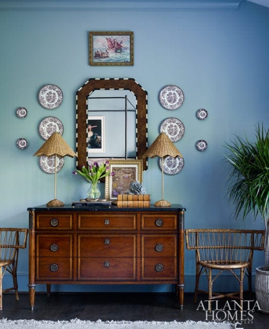 Blue walls with a collection of decorative plates and two paintings above a dark wood dresser with marble top, flanked by two wicker chairs and table lamps.