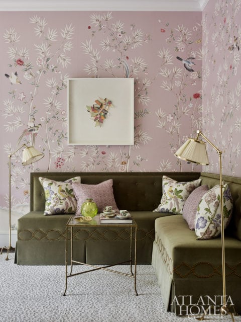 Green tufted sectional sofa with patterned pillows against a pink Chinoiserie wallpaper, a glass and brass side table with teacups, and a framed artwork.