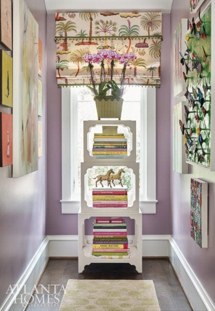 Hallway with lavender walls, featuring a window with a tropical print roman shade, a white tiered shelf with books and horse figurines, and artwork on the walls.