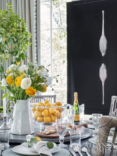 Dining table set with white plates, silverware, glassware, a bouquet of yellow and white flowers, a bowl of lemons, and a bottle of champagne. A framed artwork of a white bird hangs on the wall.