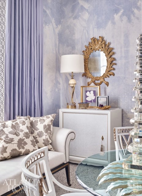 Light purple curtains frame a wall with blue and white scenic wallpaper. A white cabinet holds a gold ornate mirror, a lamp, and framed art.
