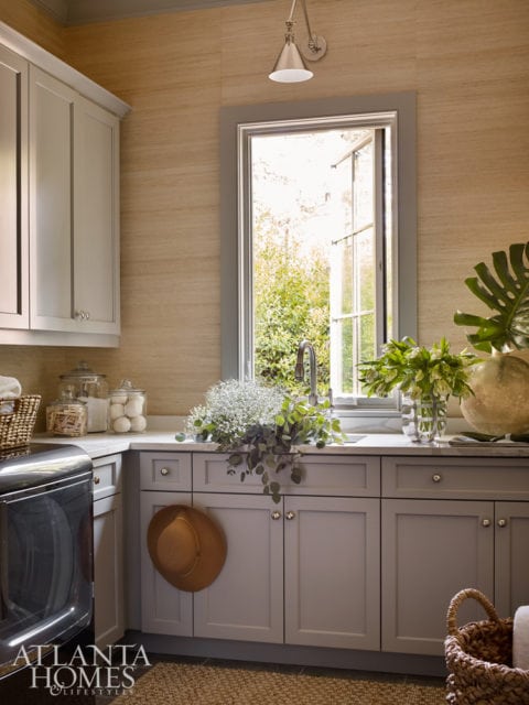 Light gray laundry room cabinets with a white countertop and a large window overlooking greenery. A straw hat hangs on one cabinet door.