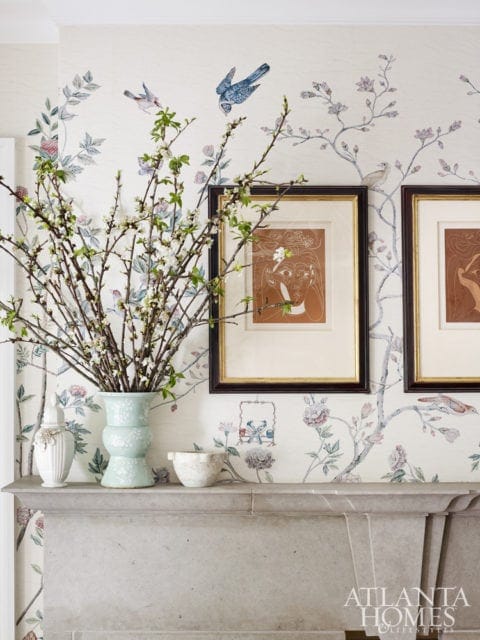 Stone fireplace mantel topped with a mint green vase of blooming branches, a white urn, and a decorative bowl, against wallpaper with a bird and floral motif.