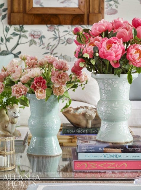 Two pale blue ceramic vases filled with pink peonies and tulips sit on a glass coffee table. Books are stacked beneath the vases.